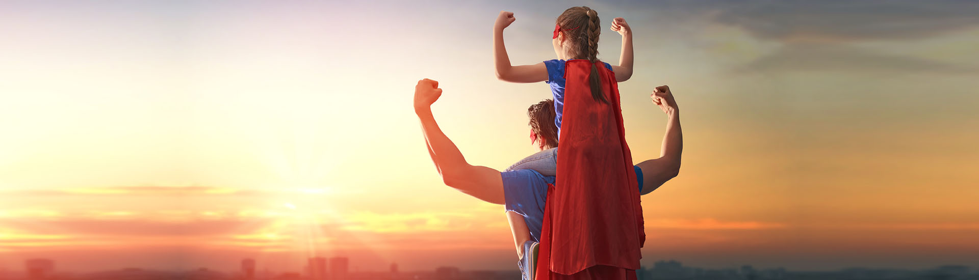 child sitting on top of parents shoulders flexing while looking at sunset